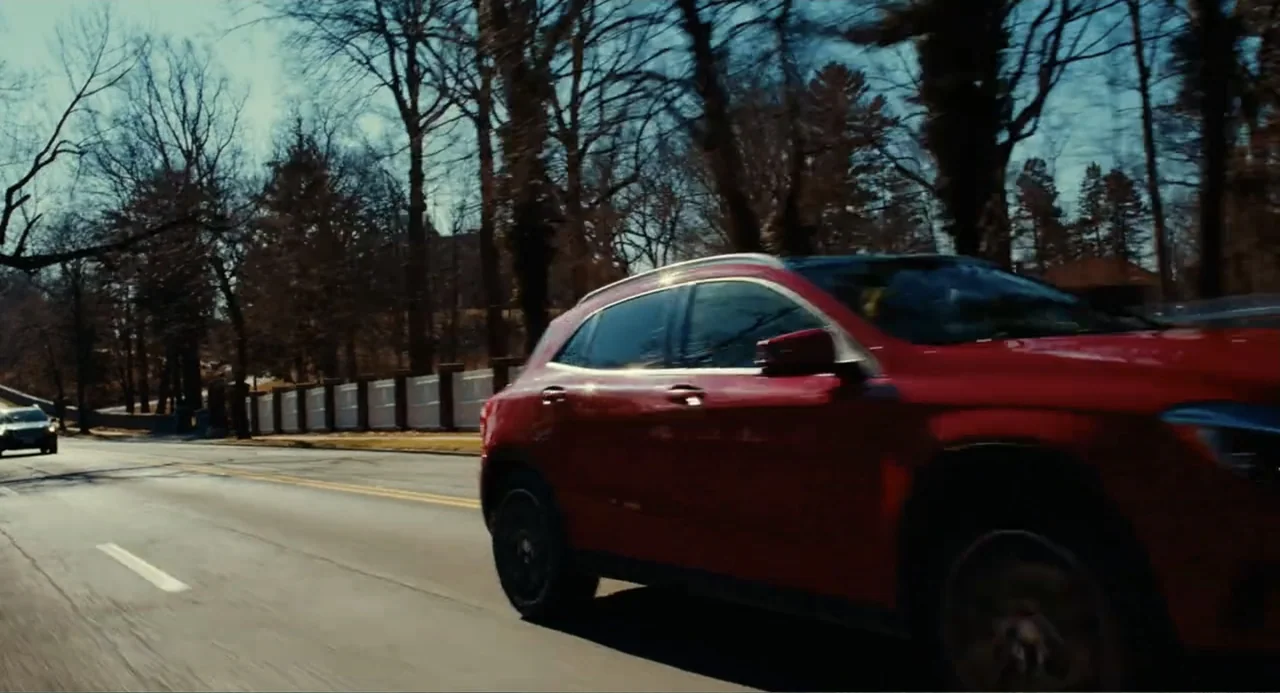 An Imperfect Murder (2017) movie still: medium — A red car drives down a paved road lined with bare trees. White fence panels follow the curve of…