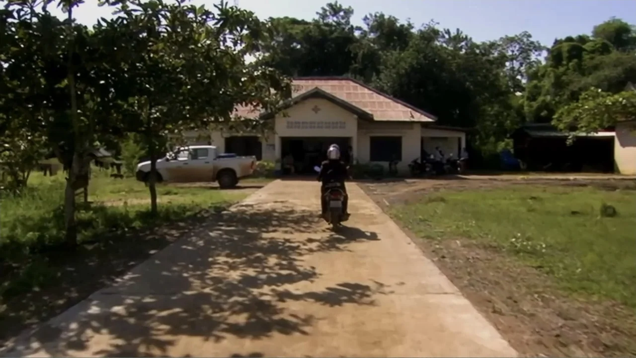 Sister (2014) movie still: wide — A rural property features a single-story white building at the end of a driveway. A person on a motorcycle drives…