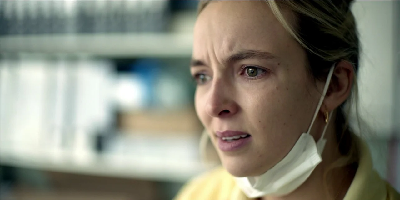 Help (2021) movie still: closeup — A workspace with blurred white shelves filled with folders in the background. A woman in a yellow shirt looks…