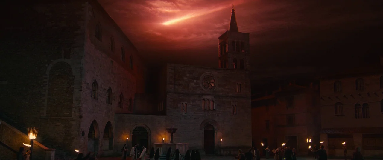Benedetta (2021) movie still: establishing — A stone town square features a Romanesque church facade under a dark red sky. A group of robed figures…