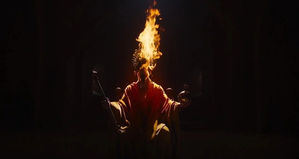 The Green Knight (2021) movie still: medium — A dark stone space with vaulted arches in the background and foreground silhouettes. A person sits on…