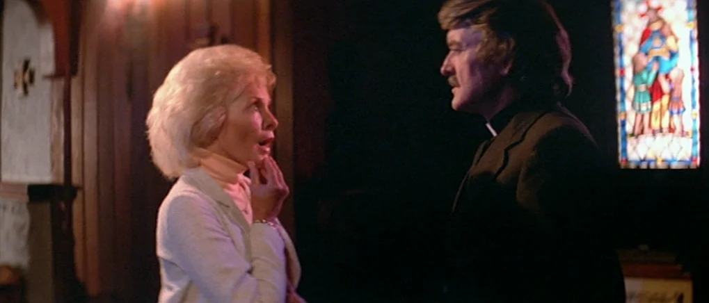 The Fog (1980) movie still: medium — A dark, wood-paneled room is dimly illuminated by a nearby stained glass window. A blonde woman speaks to a…