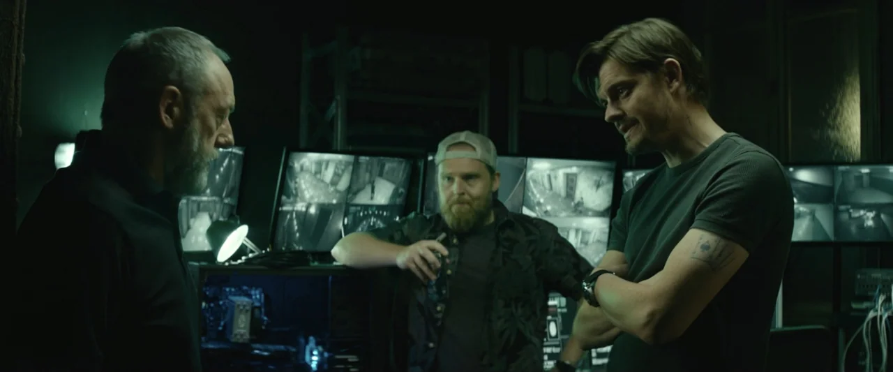 The Vault (2021) movie still: medium — A dark, cramped surveillance room filled with glowing computer monitors and technical equipment. Three men…