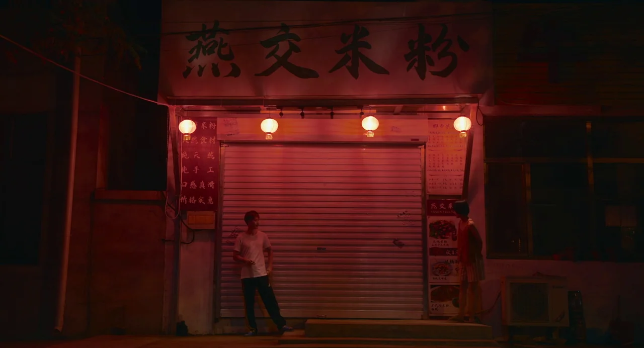 White River (2023) movie still: wide — A storefront with a closed metal rolling shutter displays Chinese characters on a lightbox. Two people stand…