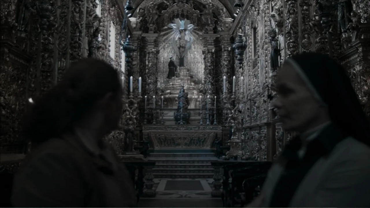 The Grotto (2022) movie still: medium — A dimly lit church interior features highly ornate gilded wood carvings surrounding the altar. Two women…