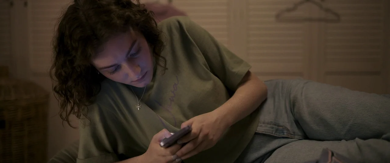 Talk To Me (2022) movie still: medium — A dark, dimly lit indoor setting features a woman resting on a surface. She holds a glowing smartphone while…