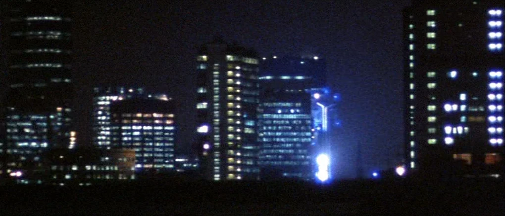 Wonderland (1999) movie still: establishing — A nighttime view of a cluster of high-rise skyscrapers with many illuminated windows. The dark sky…