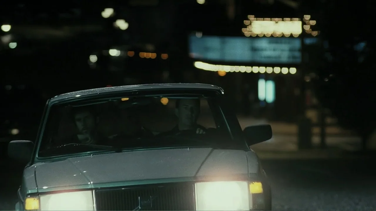 Mr. Brooks (2007) movie still: medium — A city street at night with an out-of-focus background containing a neon-lit sign. Two men sit inside the…