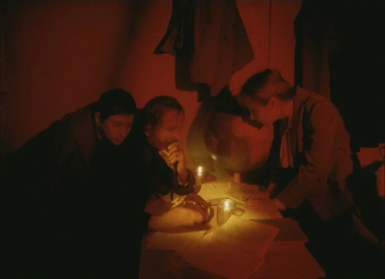 Russian Symphony (1994) movie still: medium — A dimly lit room holds three people clustered around a table at night. They examine papers together…
