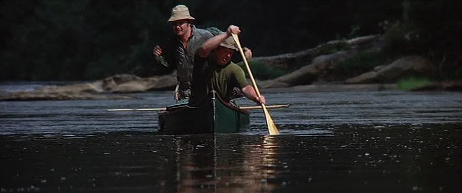 Deliverance (1972) movie still: fullbody — Two men in a dark green canoe navigate a slow-moving river in a forest. The shoreline features rocky…