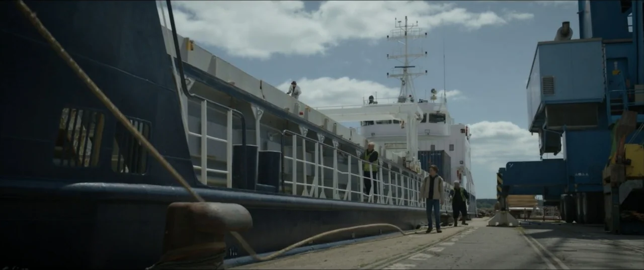 Kraven the Hunter (2024) movie still: wide — A commercial dock features a large dark ship moored alongside blue industrial cranes. A man in a beige…