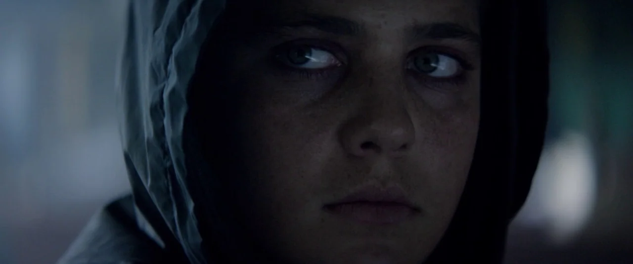 Kissing Candice (2017) movie still: closeup — A dark environment surrounds a teenage girl wearing a hood in close proximity. The subject stares off…