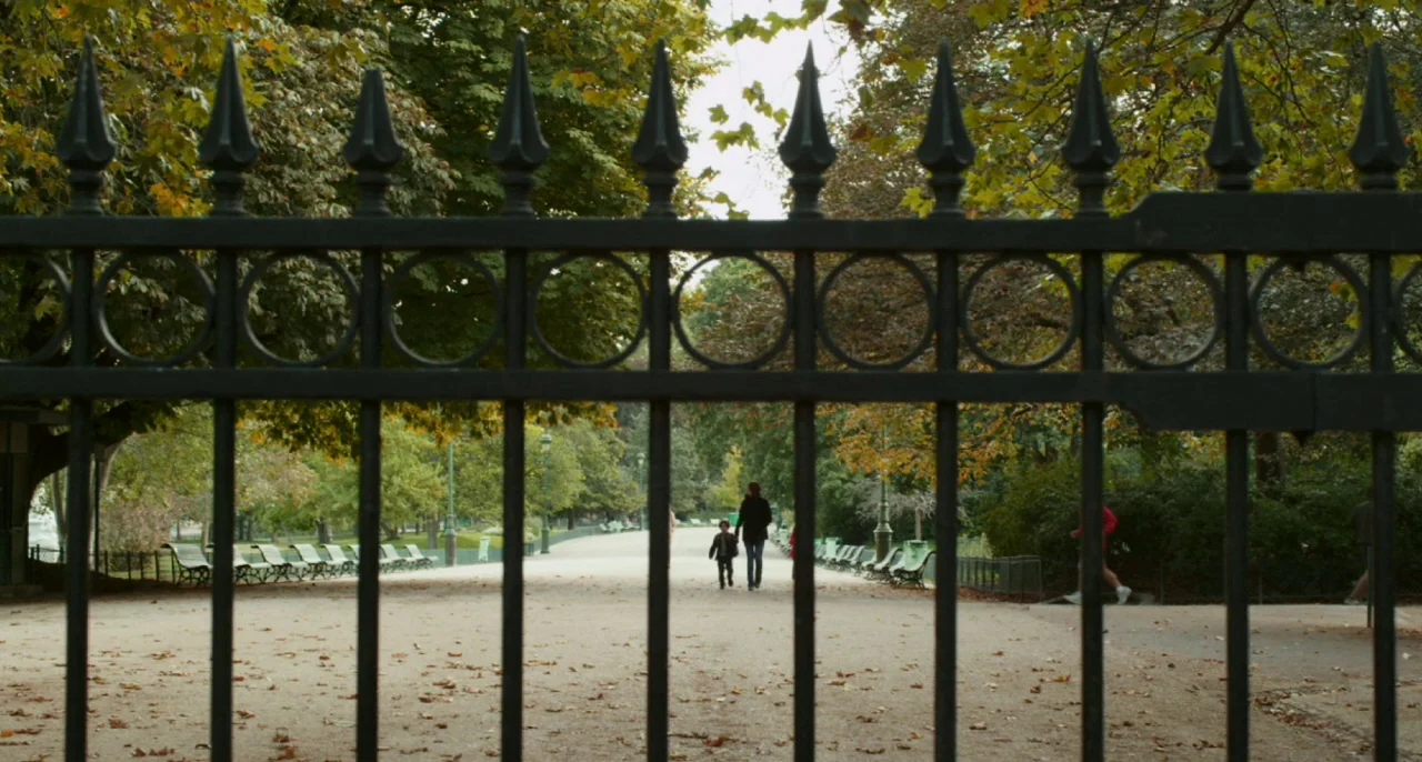 Bastards (2013) movie still: wide — A view of a public gravel walkway behind a tall, black decorative metal gate. An adult woman and a young child…