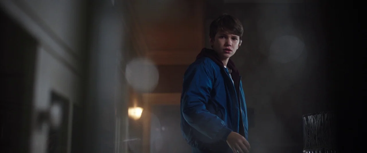 Child's Play (2019) movie still: medium — A dimly lit hallway features a wall sconce casting soft light. A teenage boy in a blue jacket looks toward…
