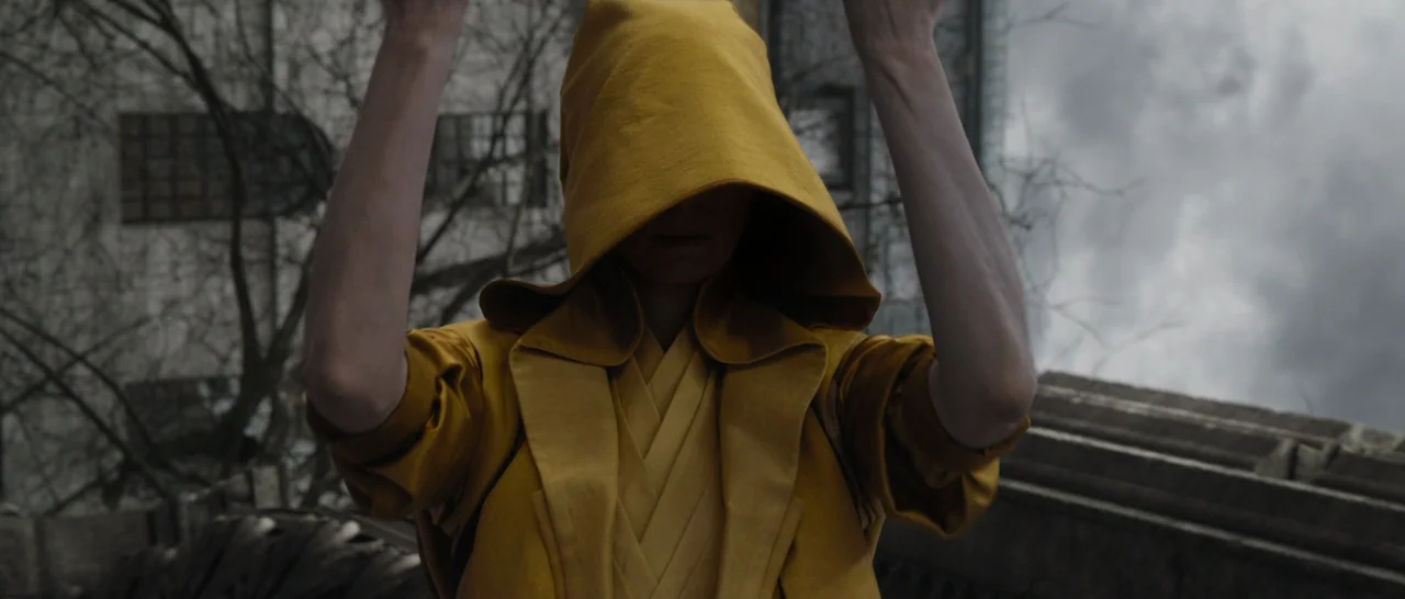 Doctor Strange (2016) movie still: medium — A person wearing a golden yellow hooded garment stands on a rooftop against a backdrop of leafless trees…