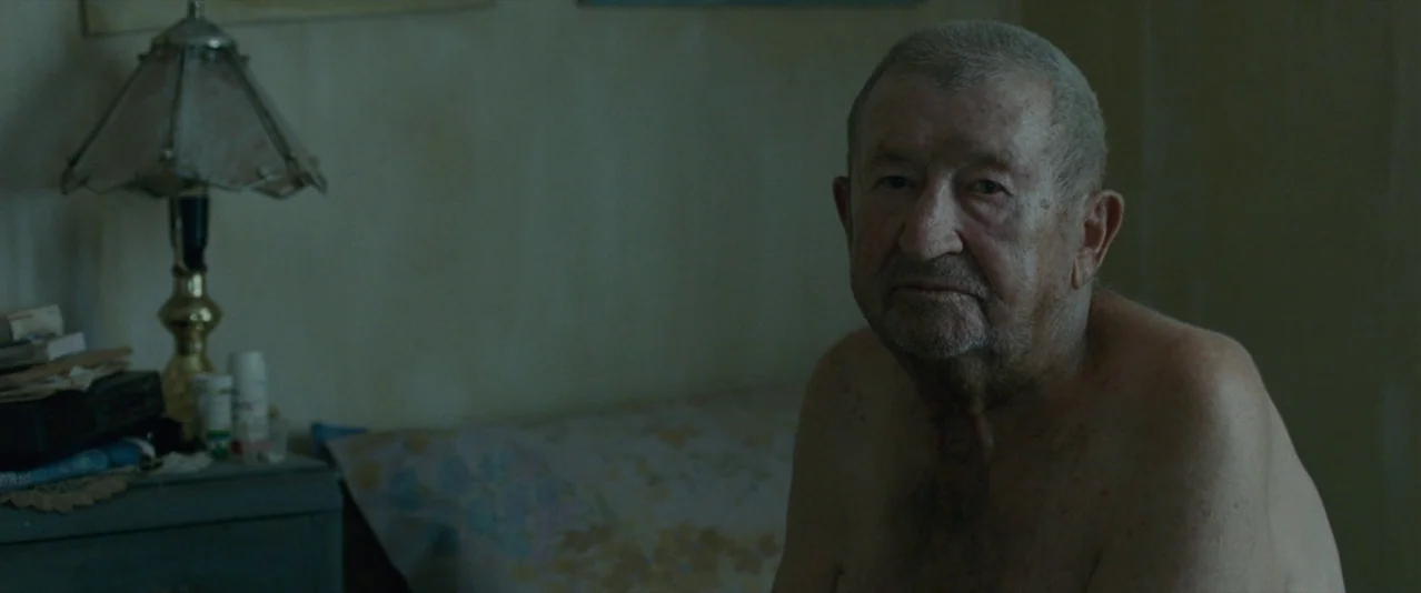 The Rover (2014) movie still: medium — A dimly lit room contains a bedside table with a lamp and medication. An older man sits shirtless on the bed…