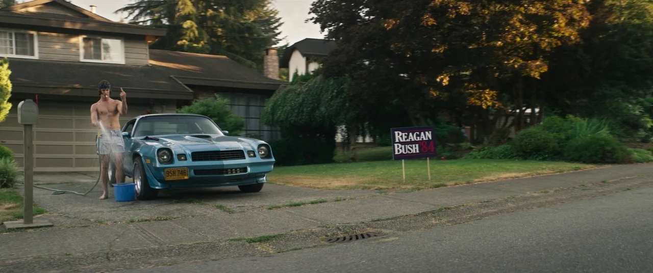 Summer of 84 (2018) movie still: wide — A quiet suburban driveway in the 1980s features a blue vintage muscle car. A young man washes the car with a…