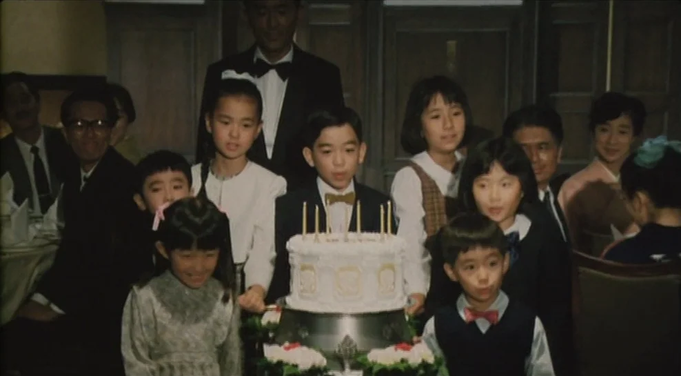Madadayo (1993) movie still: medium — A formal interior space features a group of children and adults around a table. They are positioned around a…