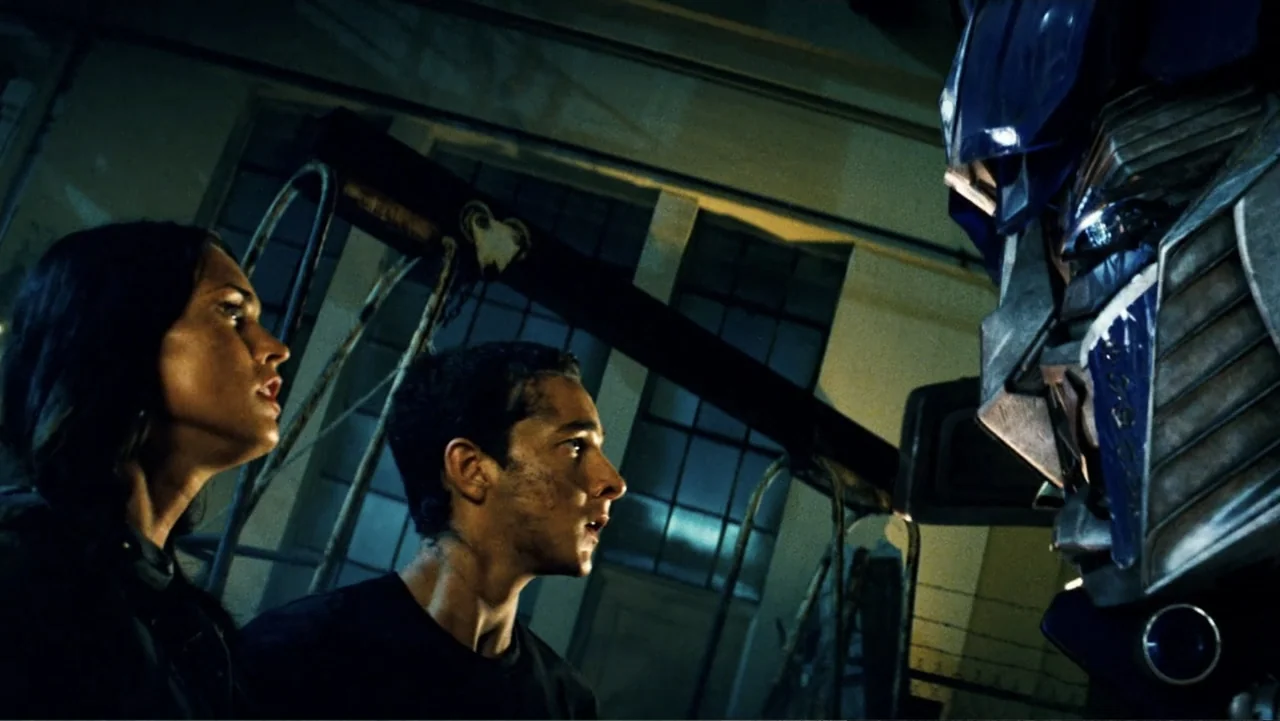 Transformers (2007) movie still: medium — An dimly lit industrial hangar interior features large metal structural supports and paneled windows. A…