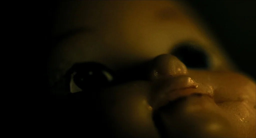 The Fall movie still: closeup — A close-up of a pale plastic doll face emerging from deep shadows. One eye is visible in the darkness while the nose…