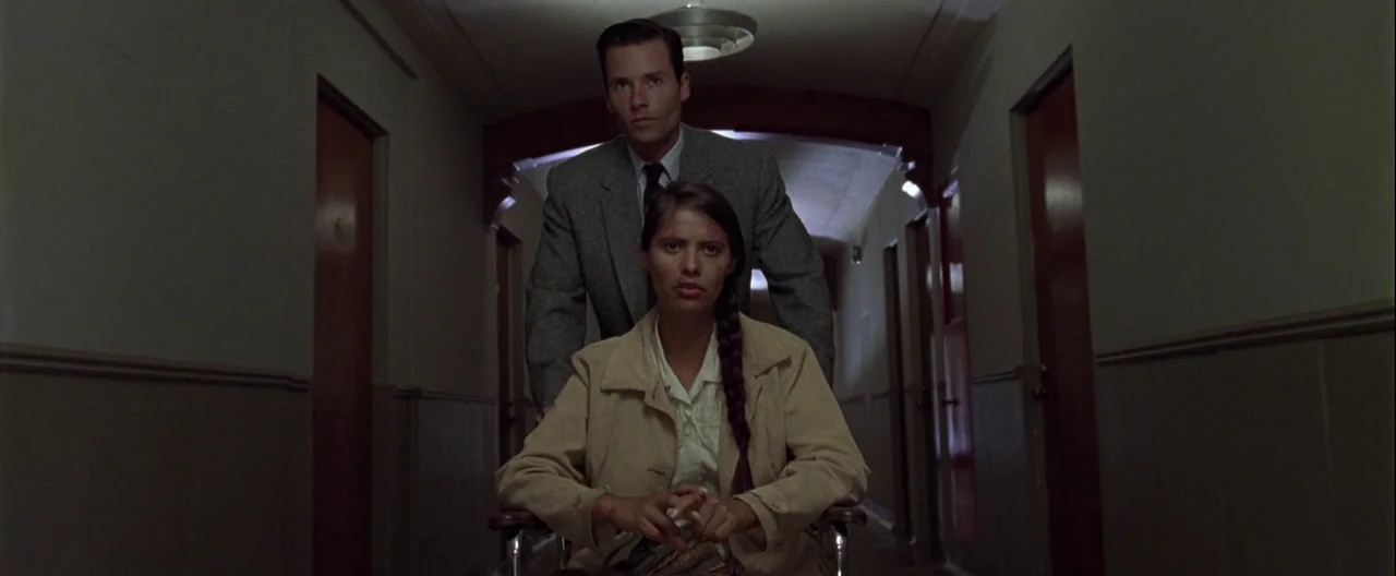 L.A. Confidential (1997) movie still: medium — A long, dimly lit building hallway features dark doors lining both sides of walls. A man in a suit…