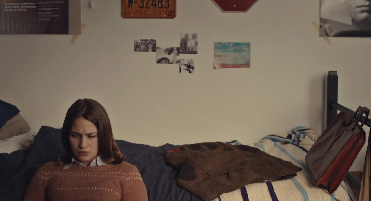 Mistress America (2015) movie still: medium — A dorm room wall decorated with vintage photographs and a New York license plate. A young woman sits…