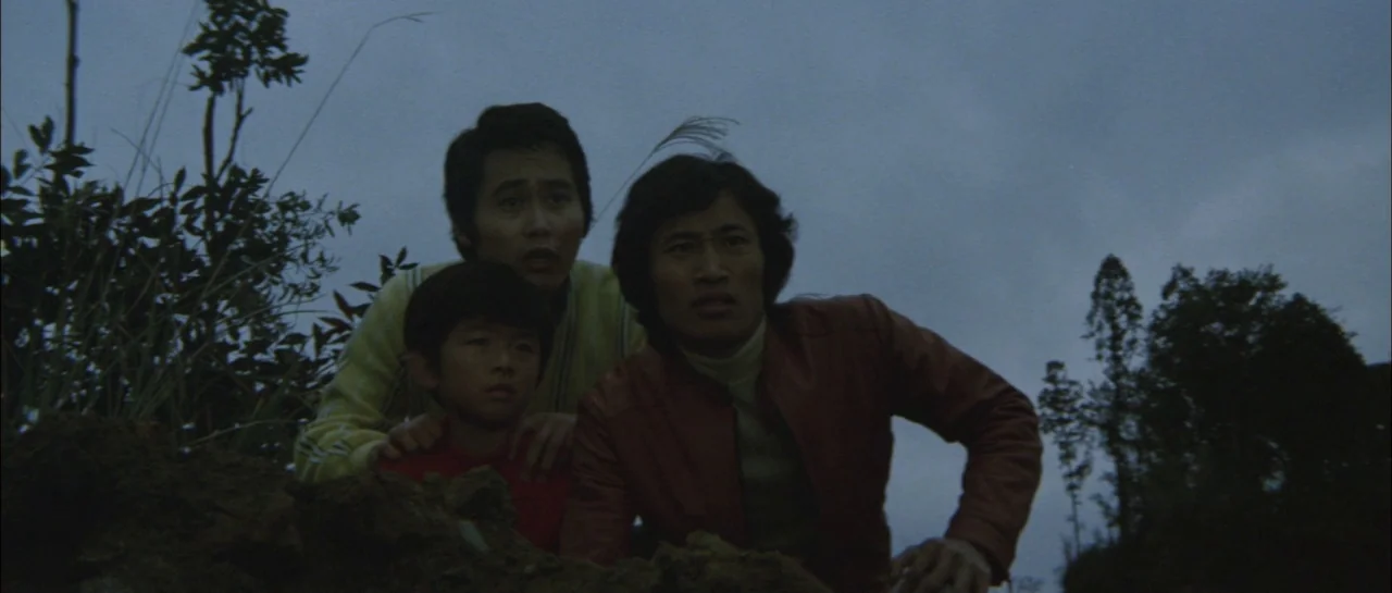 Godzilla Vs Megalon (1973) movie still: medium — An open grassy slope under an overcast twilight sky at dusk. Three males in colorful shirts crouch…