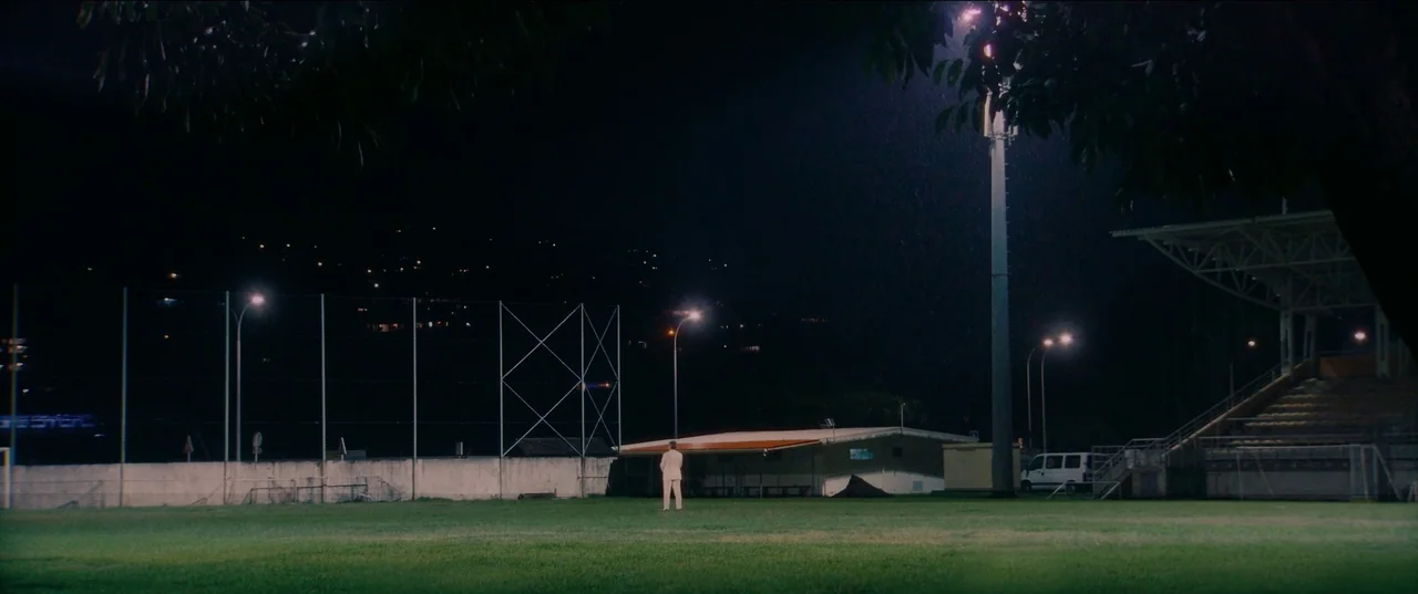 Pacifiction (2022) movie still: wide — A nighttime view of a grassy athletic field illuminated by overhead stadium lights. A person in a light suit…