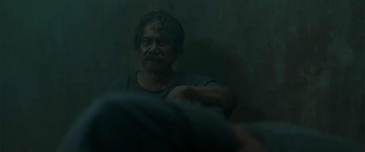 The Unholy (2022) movie still: medium — A dimly lit, windowless room features plain, textured dark walls. An older man in a gray shirt sits on the…