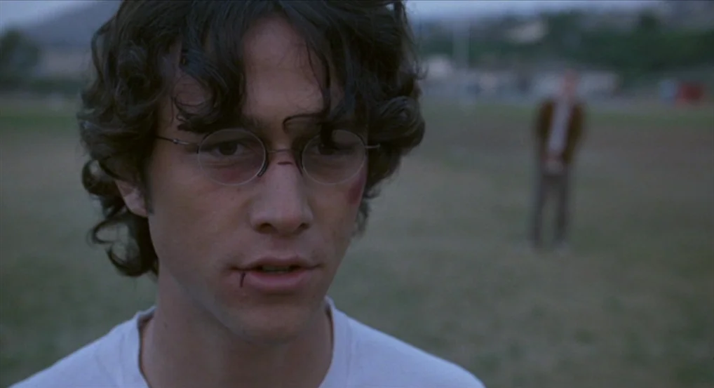 Brick (2005) movie still: closeup — A man with a bruised face stands in a grassy field at dusk. Another blurry figure stands in the background on…