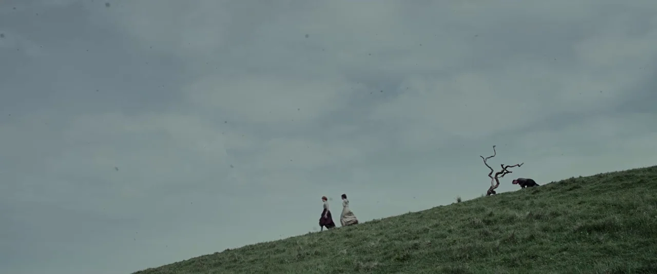 Apostle (2018) movie still: wide — A vast, overcast grassy hillside stretches toward a cloudy sky. Two women walk slowly up the incline near a lone,…