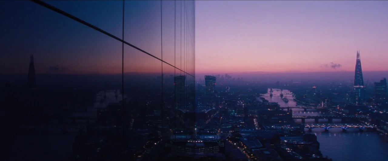 No Time To Die (2021) movie still: establishing — A dark glass skyscraper facade occupies the left side of the frame. The city of London is visible…
