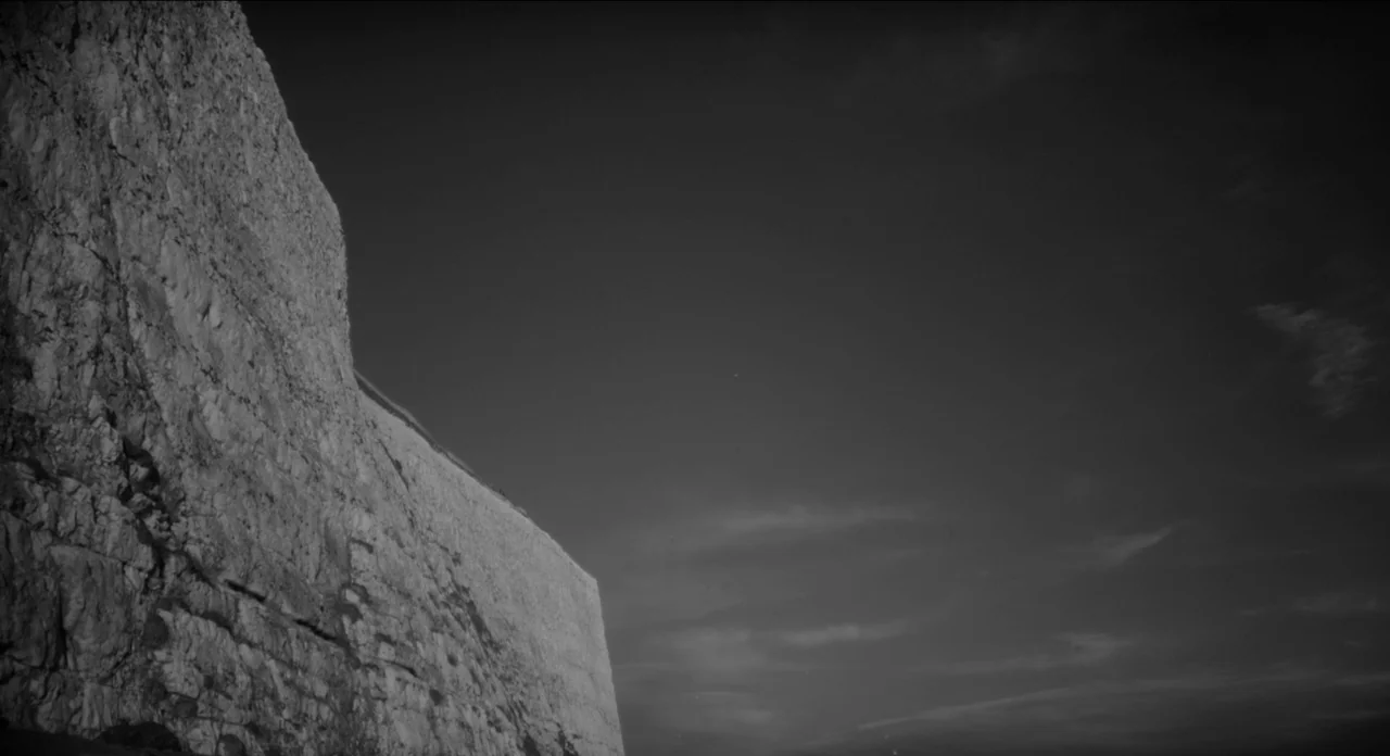 One More Time With Feeling movie still: establishing — A vertical white chalk cliff face rises sharply on the left side of frame. The right side…