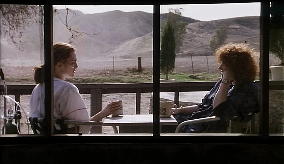 Safe movie still: medium — A wooden porch overlooks a dry, hilly rural landscape with sparse vegetation. Two women sit at a white table while…