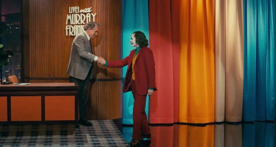Joker (2019) movie still: fullbody — A television talk show set features wood-paneled walls and vibrant multicolored vertical curtains. Two men…