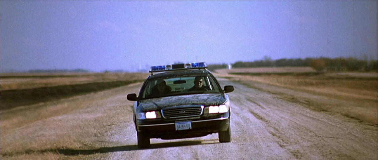 Fear X (2003) movie still: wide — A dark police cruiser travels down a long, dusty gravel road in an open field. Two men occupy the front seats of…