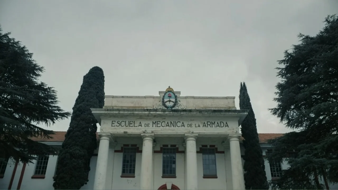 Transfers (2024) movie still: establishing — A stone neoclassical building features a large emblem and text above four main columns. Tall evergreen…