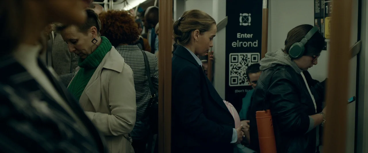 The Pod Generation (2023) movie still: medium — A crowded commuter transit car with vertical metal poles and advertisements on the wall. A woman…