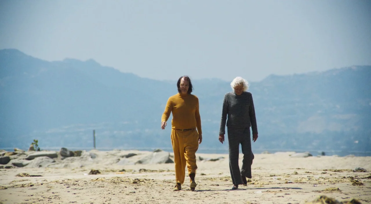 The Greasy Strangler (2016) movie still: fullbody — A wide, flat beach expanse is flanked by distant blue mountain silhouettes. Two men in distinct…