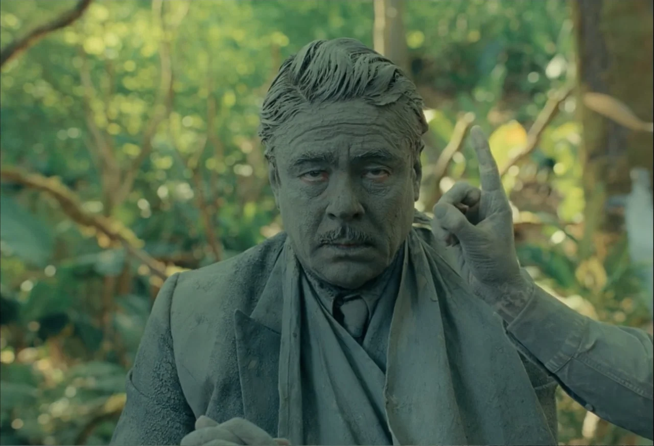The Phoenician Scheme (2025) movie still: medium — A man in a mud-covered suit stands before a backdrop of thick green vegetation. The subject…