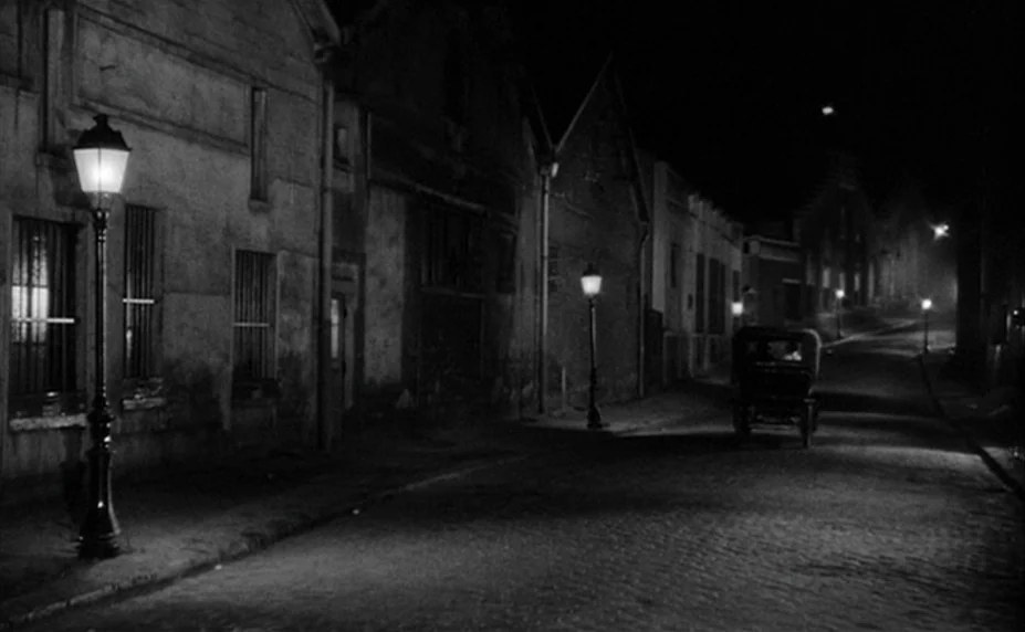 Judex (1966) movie still: establishing — A narrow cobblestone street is lined with weathered stone buildings and iron-post streetlamps at night. A…