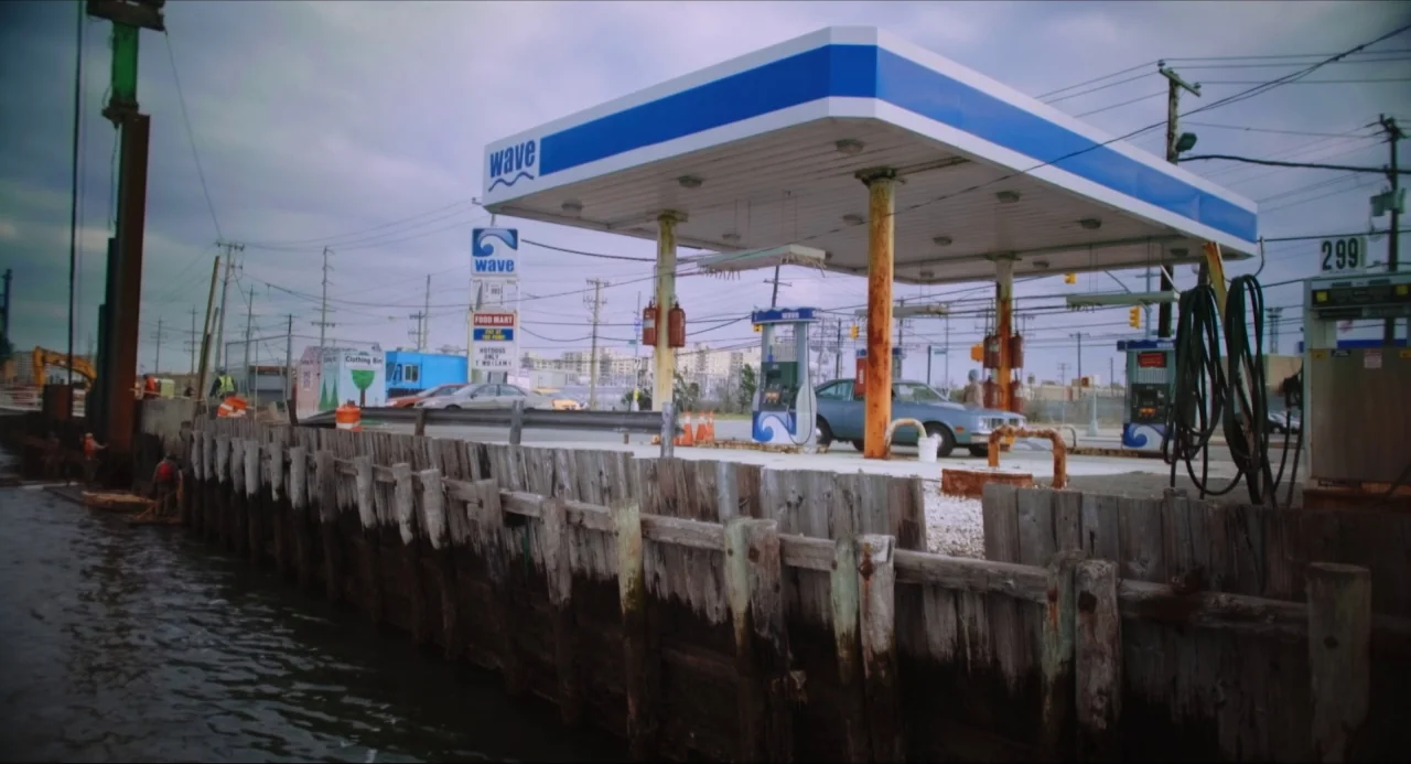 Like Me movie still: wide — A waterfront gas station sits adjacent to a weathered wooden bulkhead on the shore. A blue fuel canopy provides cover…