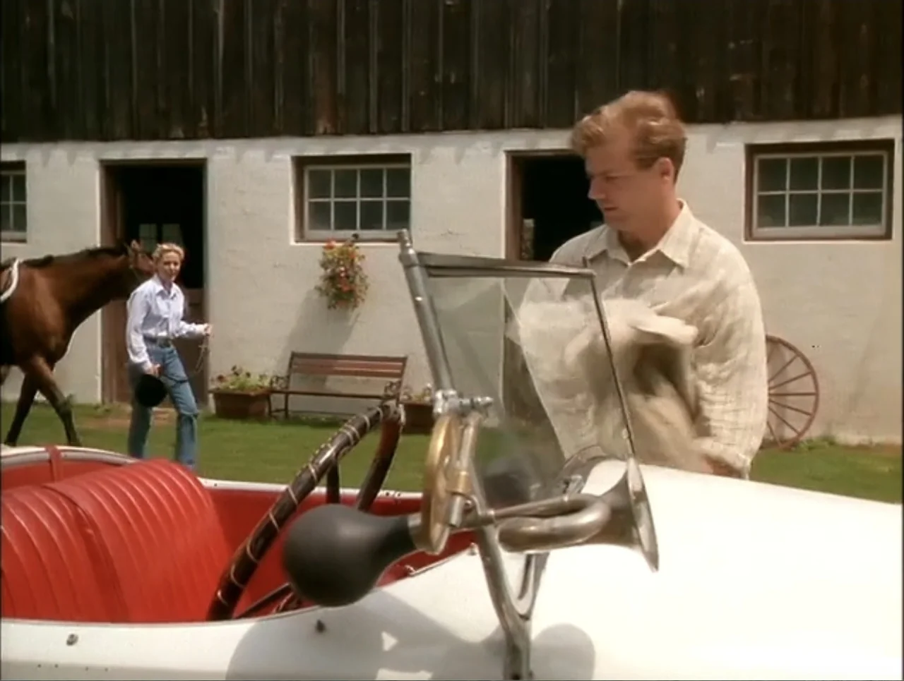 At the Midnight Hour (1995) movie still: medium — A man stands by a vintage open-top car with red interior in front of a white barn. A woman walks…