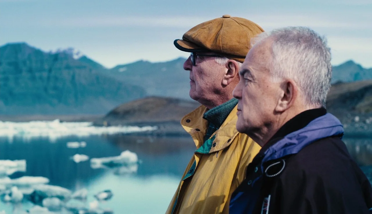 Land Ho! (2014) movie still: medium — Two men stand at the edge of a glacier lagoon surrounded by icy water. The background features distant…