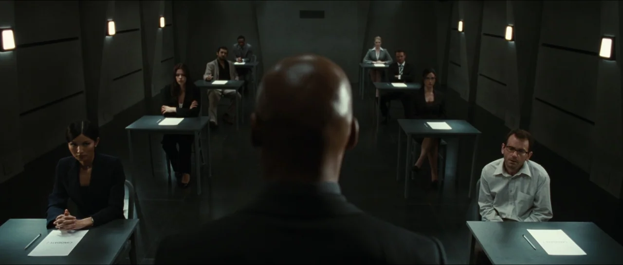 Slackers (2002) movie still: medium — A stark, windowless examination room contains multiple rows of individual desks for test participants. Eight…