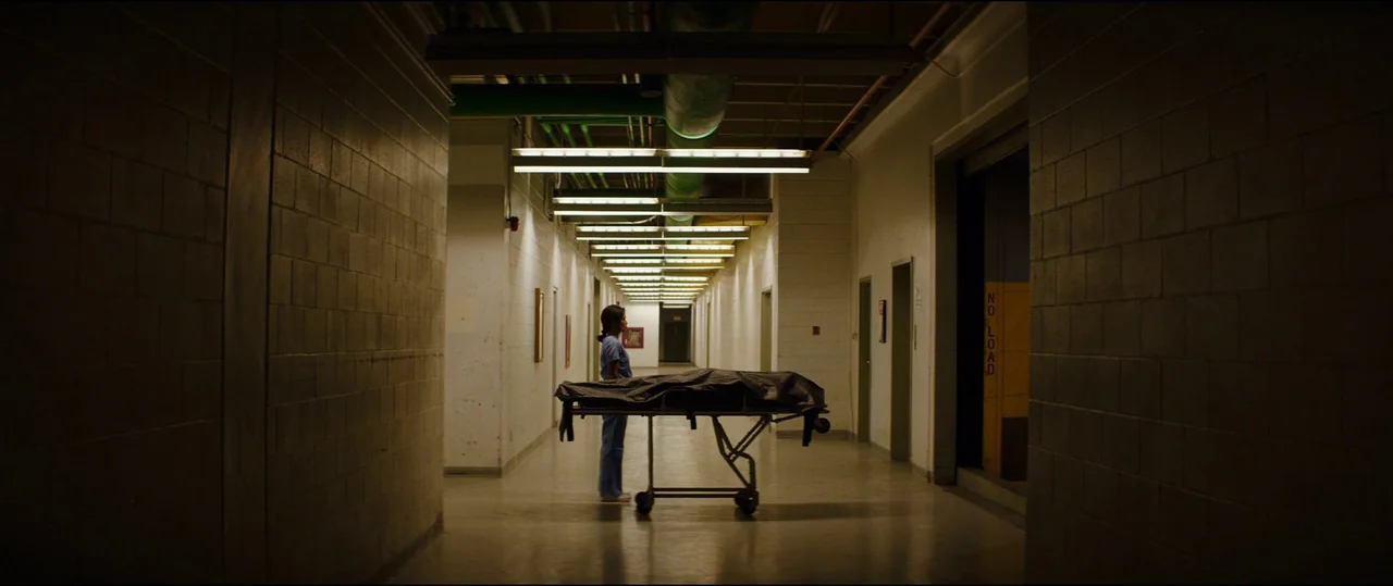 El Chicano (2018) movie still: wide — A long, industrial hospital corridor features white cinderblock walls and exposed overhead pipes. A lone…