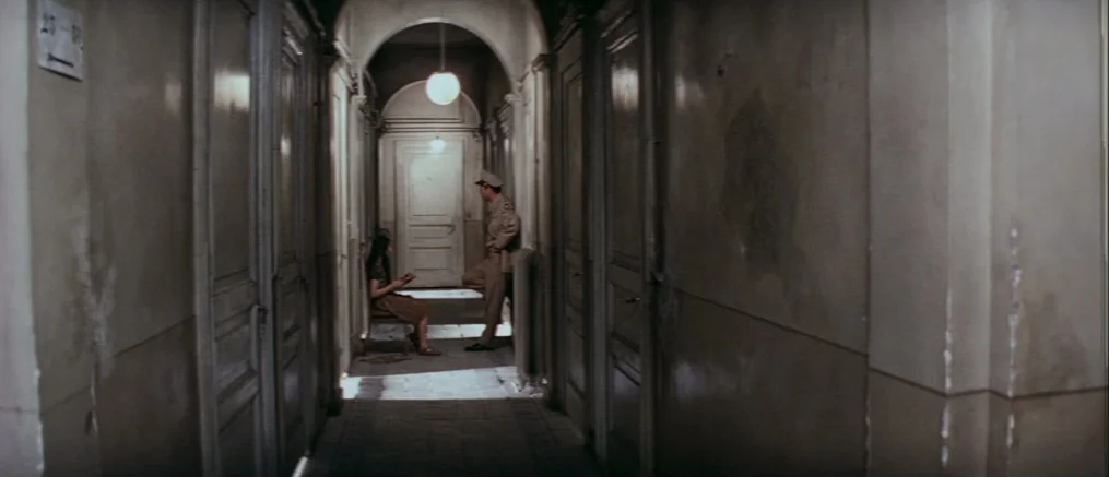 Catch 22 (1970) movie still: wide — A long, narrow apartment hallway features tall wooden doors and dim, aged walls. A seated woman and a standing…