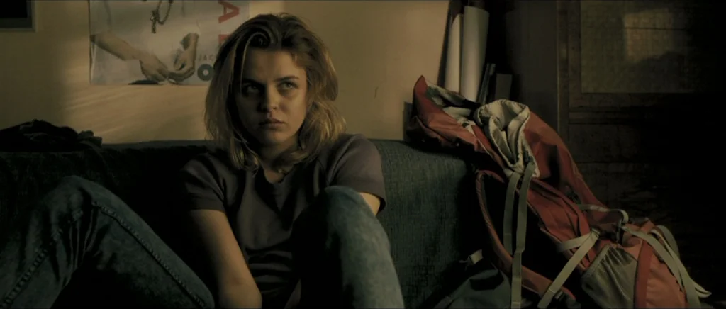 Lasting (Nieulotne) (2013) movie still: medium — A dark room with a sofa sits before a wall decorated with a poster. A young woman with blonde hair…