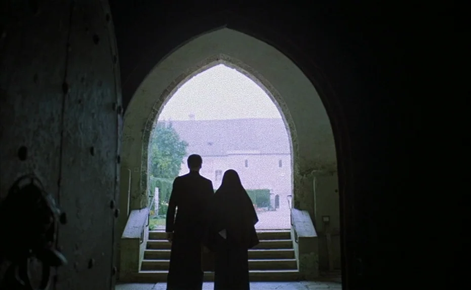 To The Devil a Daughter (1976) movie still: wide — A stone archway frame provides a view of a light-colored building exterior. A man and a woman in…
