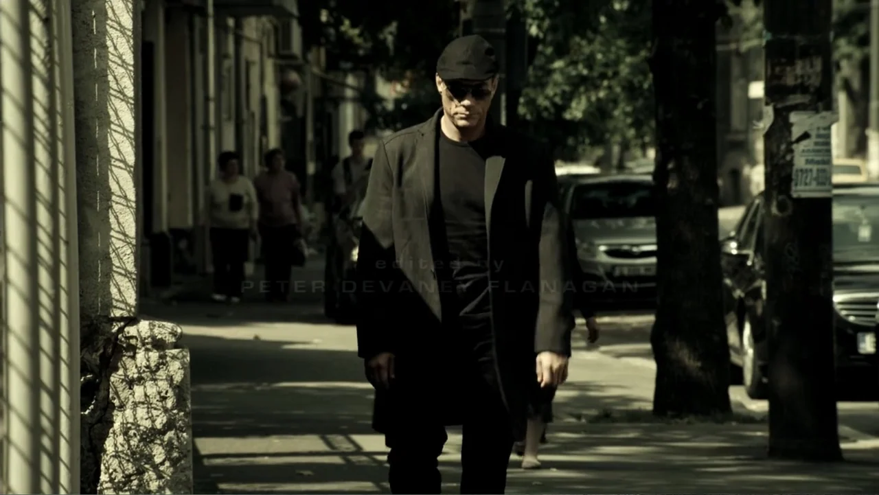 Assassination Games (2011) movie still: fullbody — An urban sidewalk runs along a row of buildings under bright daylight conditions. A man in a dark…